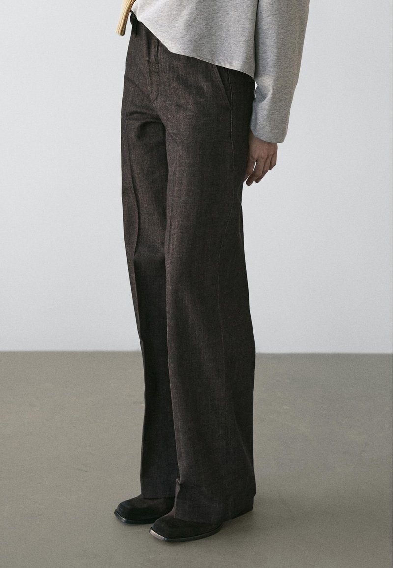 Persoon die donkere broek met wijde pijpen en zwarte schoenen draagt, met een lichtgrijs shirt met lange mouwen, staand op een grijze vloer tegen een effen achtergrond.