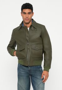 Giovane uomo che indossa una giacca bomber in pelle verde oliva con tasche anteriori e jeans blu, in piedi su uno sfondo bianco.