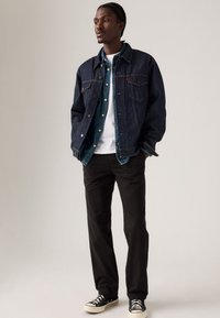Donkerdenim jacket over een blauwe shirt, witte t-shirt, zwarte broek, en zwarte sneakers met een witte zool, geplaatst tegen een lichte achtergrond.