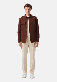 Braune Jacke mit Knöpfen und zwei Brusttaschen, getragen über einem cremefarbenen Oberteil und beigen Hosen, ergänzt durch weiße Sneakers. Einfaches Design und Passform.