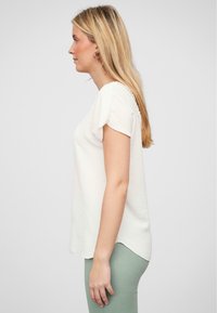 Hvit kortermet topp med teksturert stoff og detaljering på siden, kombinert med lysegrønne leggings. Rett snitt, avslappet passform.