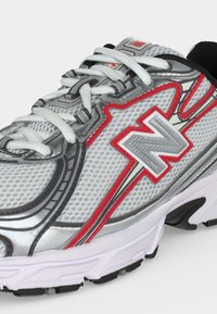 Κοντινό πλάνο γκρι και άσπρου παπουτσιού τρεξίματος New Balance με κόκκινες λεπτομέρειες, πλέγμα στο πάνω μέρος, άσπρα κορδόνια και μαλακή σόλα.