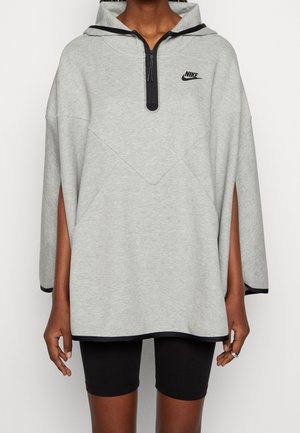 Person iført en lysegrå Nike hættetrøje med sort lynlås og kant, kombineret med sorte cykelshorts, stående foran en hvid baggrund.