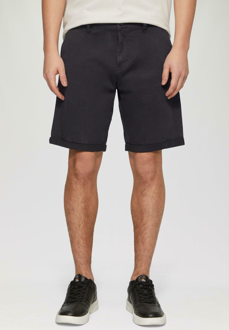 Shorts noirs en coton avec ourlets roulés, comportant des poches latérales et une coupe droite. Portés avec des baskets noires et une chemise de couleur claire.