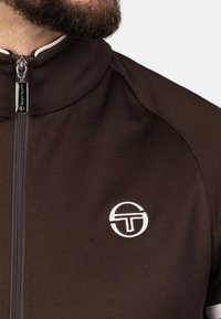 Veste zippée marron en tissu lisse avec un col haut. Présente un logo blanc sur la poitrine et une fermeture éclair métallique.