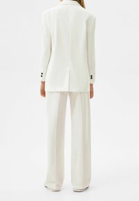 Kvinna klädd i en vit skräddarsydd blazer och matchande vida byxor, stående med ryggen mot kameran mot en vit bakgrund.