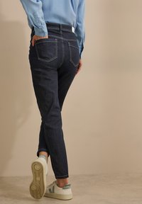 Jean skinny en denim foncé avec taille haute, coutures visibles et deux poches arrière. Associé à des baskets blanches avec des accents gris.