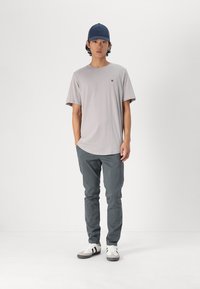 Camiseta de manga corta gris claro con un pequeño logo, combinada con pantalones gris oscuro y zapatillas blancas con rayas negras, además de una gorra azul marino.