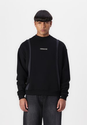 Sweatshirt noir à col montant, arborant un logo "FRANCHISE". Comprend des accents gris verticaux sur les côtés. Porté avec un pantalon gris foncé.