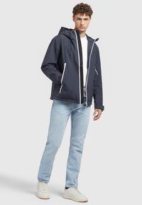 Veste imperméable marine avec fermeture éclair sur le devant, capuche ajustable, accents blancs et poches zippées, portée avec un jean bleu clair et des baskets blanches.