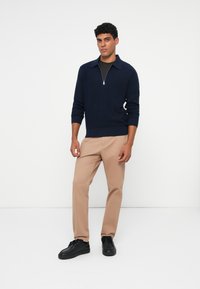 Pulover din tricot navy cu guler cu jumătate de fermoar, asortat cu pantaloni bej și adidași negri. Material texturat, design ajustat și stil minimalist.