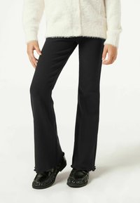 Pantalones acampanados negros con textura acanalada, con un dobladillo con volantes. Combinados con zapatos estilo mocasín negros.