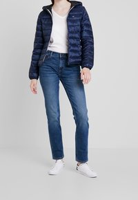 Marineblå pufferjakke med horisontale syninger, lynlåslukning og hætte. Båret med en hvid V-hals t-shirt, blå jeans og hvide sneakers.