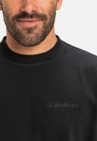 Sudadera negra de algodón, con cuello redondo y logo bordado de "Quiksilver" en gris oscuro en el pecho. Textura suave.