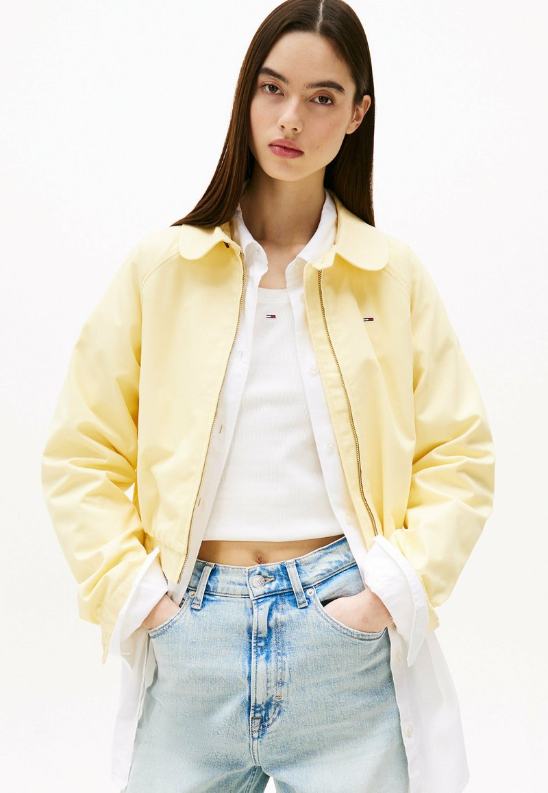 Geel cropped jasje met een kraag, ritssluiting en zijzakken. G gedragen over een witte top en gecombineerd met lichtblauwe high-waisted jeans.