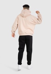 Hoodie rose poussiéreux avec un grand logo dans le dos, des poignets et un ourlet côtelés, associé à un pantalon de jogging noir et des baskets blanches, vu de derrière.