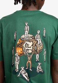 Green cotton t-shirt featuring a graphic print of a man with a beard, surrounded by smaller figures in various poses, outlined in contrasting colors.