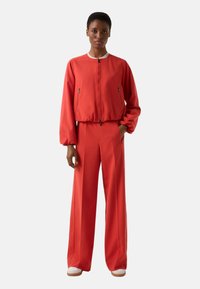Rød bomberjakke med lynlås foran, manchetter ved ærmerne og to lynlåse i siderne, parret med højtaljede, vidde bukser i matchende stof.