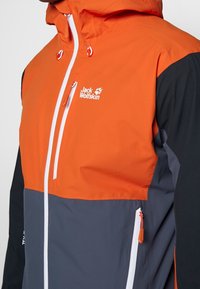 Oranje en grijze jas met capuchon, witte rits en logo. Gemaakt van duurzaam materiaal met contrasterende kleurenblokken en ritszakken.