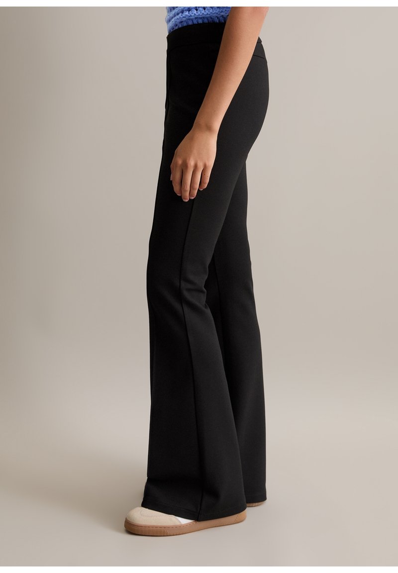 Personne portant un pantalon flare noir ajusté et des chaussures beiges, debout de profil devant un fond neutre uni.