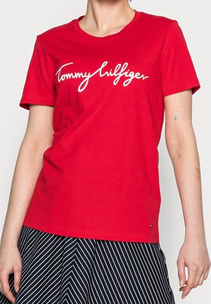 Osoba nosząca czerwoną koszulkę z krótkim rękawem z białym napisem "Tommy Hilfiger" oraz czarno-białą spódnicę w paski.