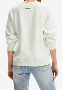 Felpa bianca con scollatura a girocollo, polsini e orlo a costine, caratterizzata da un piccolo logo nero "Desigual." sulla parte alta della schiena. Design casual.