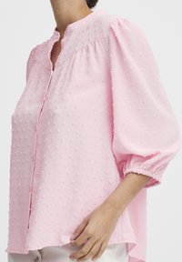 Blusa rosa chiaro con un motivo a pois testurizzato, scollatura rotonda e maniche a sbuffo, con polsini arricciati e chiusura con bottoni.