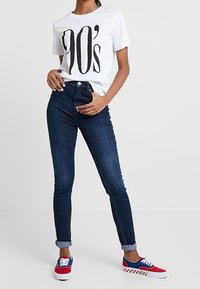 Wit T-shirt met een grote zwarte "90's" opdruk, donkerblauwe hoge taille jeans en rood-wit-blauw patroon sneakers met veters.