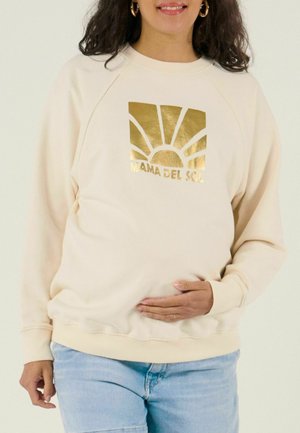 Femme portant un sweat crème avec un graphisme de soleil doré et le texte "MAMA DEL SOL", tenant doucement son ventre de femme enceinte.