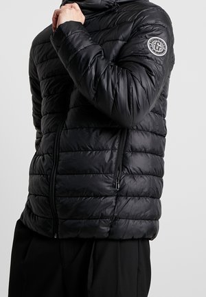 Muž v černé prošívané bundě se zipovými kapsami a kulatou nášivkou s logem na horní levé rukávu.