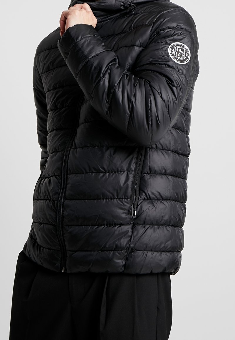 Homme portant une veste matelassée noire avec des poches zippées et un écusson rond avec logo sur la partie supérieure de la manche gauche.