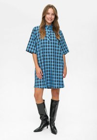 Giovane donna in un vestito a maniche corte blu e nero a quadri lungo fino al ginocchio e stivali alti neri fino al ginocchio, in piedi e sorridente su uno sfondo bianco.