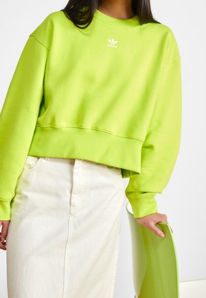 Kobieta ubrana w jaskrawo limonkową bluzę Adidas i białą spódnicę, trzymająca prawą ręką oparcie krzesła w tym samym kolorze limonkowym.