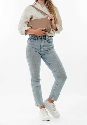 Kvinde iført lysebeige bluse og lyseblå jeans, der holder en taupe taske med hvide sneakers på hvid baggrund.