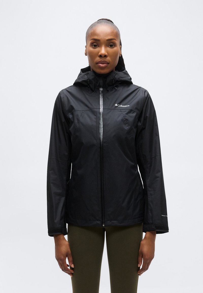 Γυναίκα που φοράει μαύρο αδιάβροχο μπουφάν Columbia με κουκούλα και φερμουάρ στο μπροστινό μέρος, στεκόμενη μπροστά σε μονόχρωμο ανοιχτό γκρι φόντο.