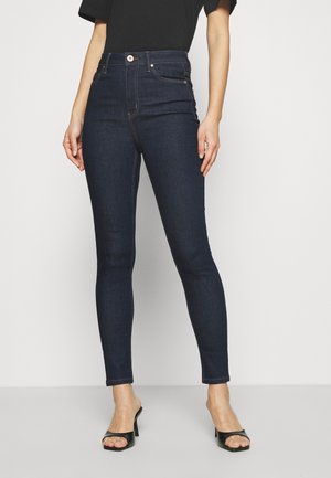 Kvinde iført mørkeblå højtaljede skinny jeans og sorte åbne hæle-sandaler, stående mod en ensartet lys baggrund.