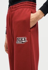 Κόκκινα φλις παντελόνια φόρμας με ελαστική μέση, πλαϊνή τσέπη και κεντημένο λογ ότυπο "NIKE CLUB" σε μαύρο και λευκό στον μηρό.