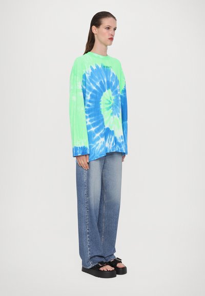 Long-sleeve tie-dye shirt in bright green and blue, paired with light blue denim jeans and chunky black sandals. Relaxed fit and soft texture.