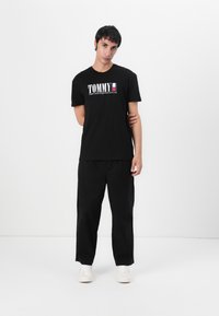 Svart bomulls T-skjorte med "TOMMY" logo i hvitt og rødt, sammen med løse svarte bukser og hvite joggesko på en nøytral bakgrunn.