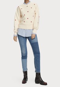 Sweatshirt crème avec broderie d'araignée rouge, assorti à une chemise rayée bleue et blanche, et un jean en denim réparé. Des bottes noires à la cheville complètent le look.