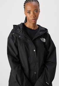 Veste imperméable noire en tissu texturé avec capuche, fermeture à boutons-pression et logo "The North Face" sur la poitrine.