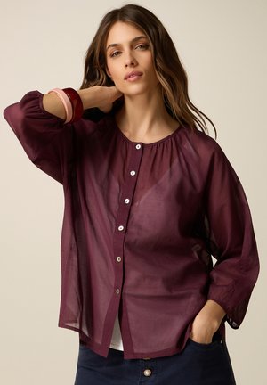 Donna che indossa una camicetta bordeaux trasparente con bottoni e maniche a sbuffo, pantaloni scuri e due braccialetti al polso, posando con una mano dietro il collo.