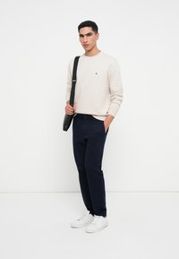 Maglione beige chiaro, pantaloni blu scuro e sneaker bianche. Il modello tiene una borsa a tracolla nera, con un piccolo logo sul maglione.