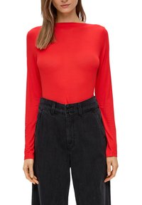 Rotes Langarmoberteil mit Rundhalsausschnitt, aus einem glatten Stoff, kombiniert mit hoch-taillierten, dunkel-schwarzen Jeans mit Doppelknopfverschluss.