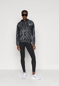 Giacca a vento a righe nere e grigie con cappuccio, abbinata a leggings neri e scarpe da ginnastica bianche. Il modello indossa occhiali da sole neri oversize.
