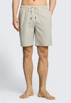 Mann trägt beige gestreifte Shorts mit Kordelzug, steht barfuß mit den Händen in den Taschen vor schlichter Hintergrund.