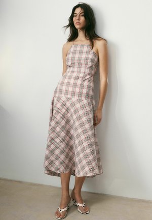 Nő ujjatlan, kockás midi ruhában, négyzet alakú nyakkivágással, mezítláb áll egy egyszerű, fehér fal előtt beltéren.