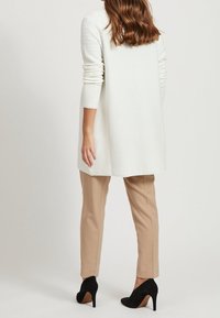 Vit ribbad långärmad cardigan, kombinerad med skräddarsydda beige byxor och svarta högklackade skor, sedd bakifrån.