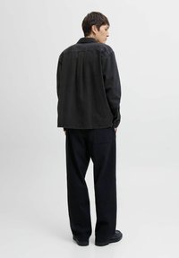 Person standing facing sideways and back, wearing a black oversized denim jacket, loose black pants, and black shoes on a plain background.