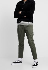 Svart manchesterjacka, randig skjorta, gröna avsmalnande byxor, svarta och vita sneakers med vita sulor. Stående pose, armarna korsade.